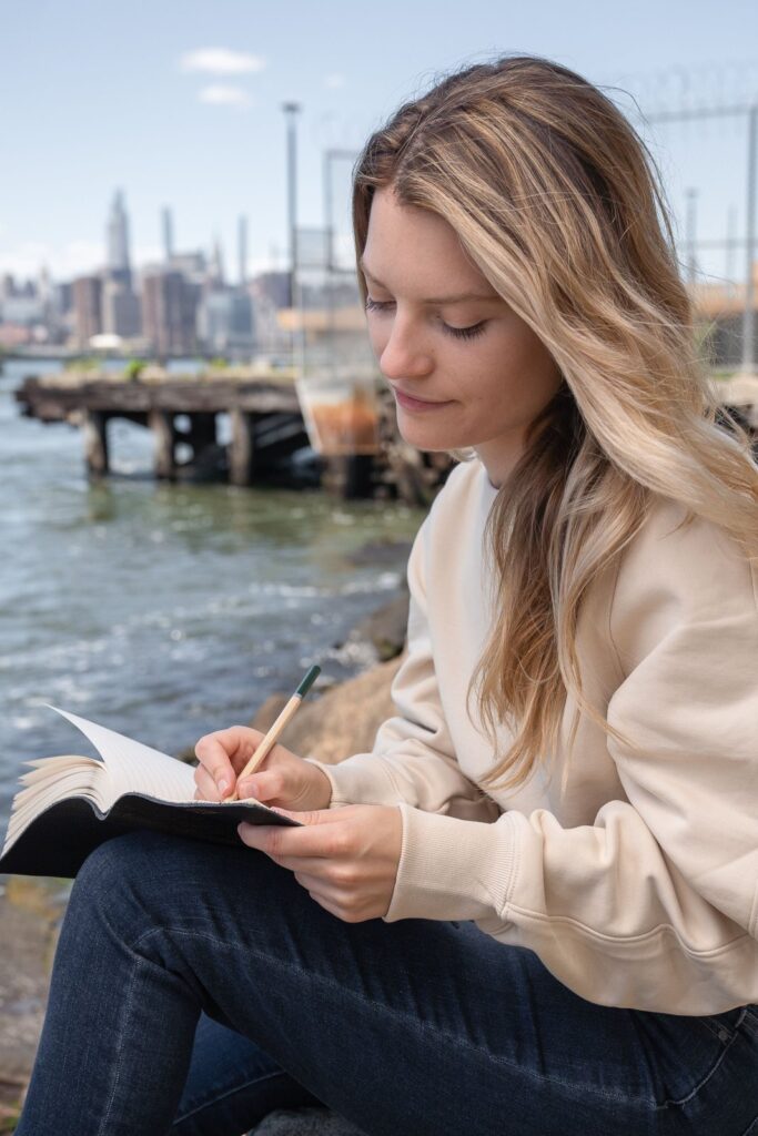 A young woman sitting by the water, writing a list in her notebook