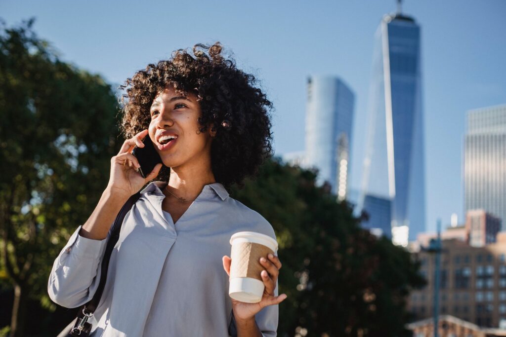A smiling woman talking on a smartphone and holding a coffee cup outdoors with the New York City skyline behind her