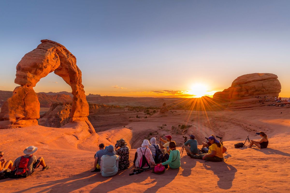 A group of friends sitting together below a red rock arch at sunset