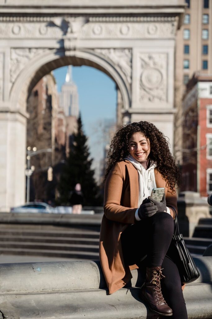 A smiling woman sitting by a New York City arch, checking her phone as she sets up how to automate savings