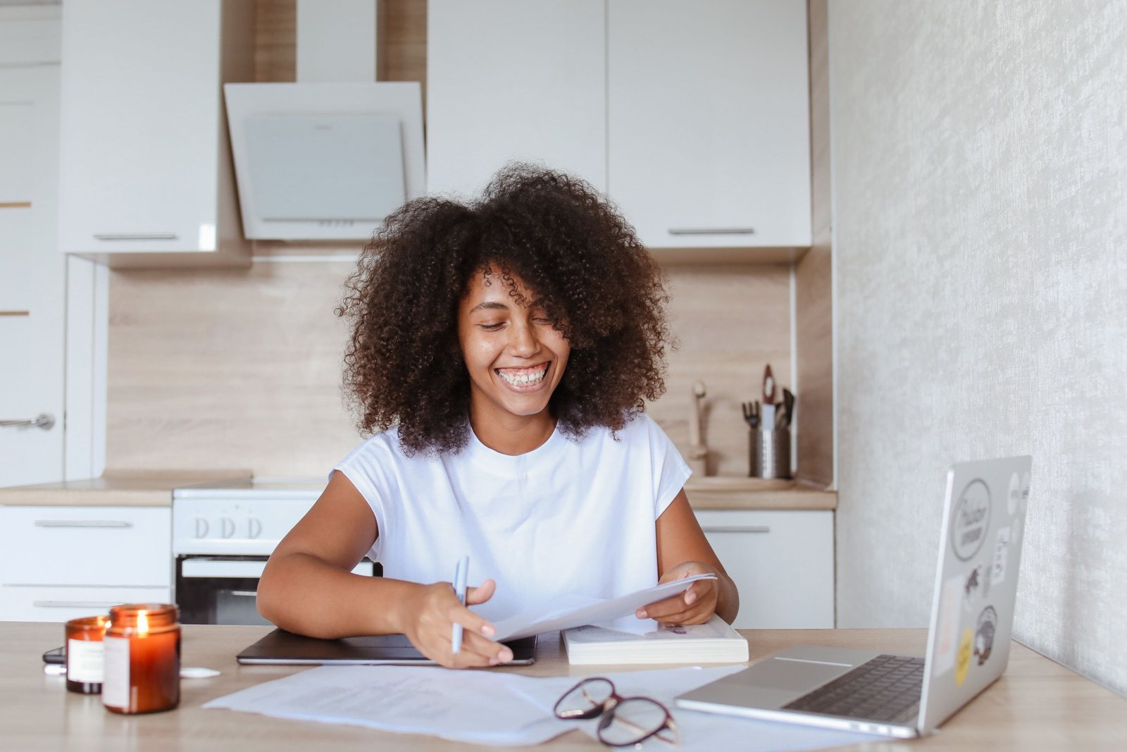 A smiling woman at her kitchen table reading a letter, with paperwork and a laptop beside her