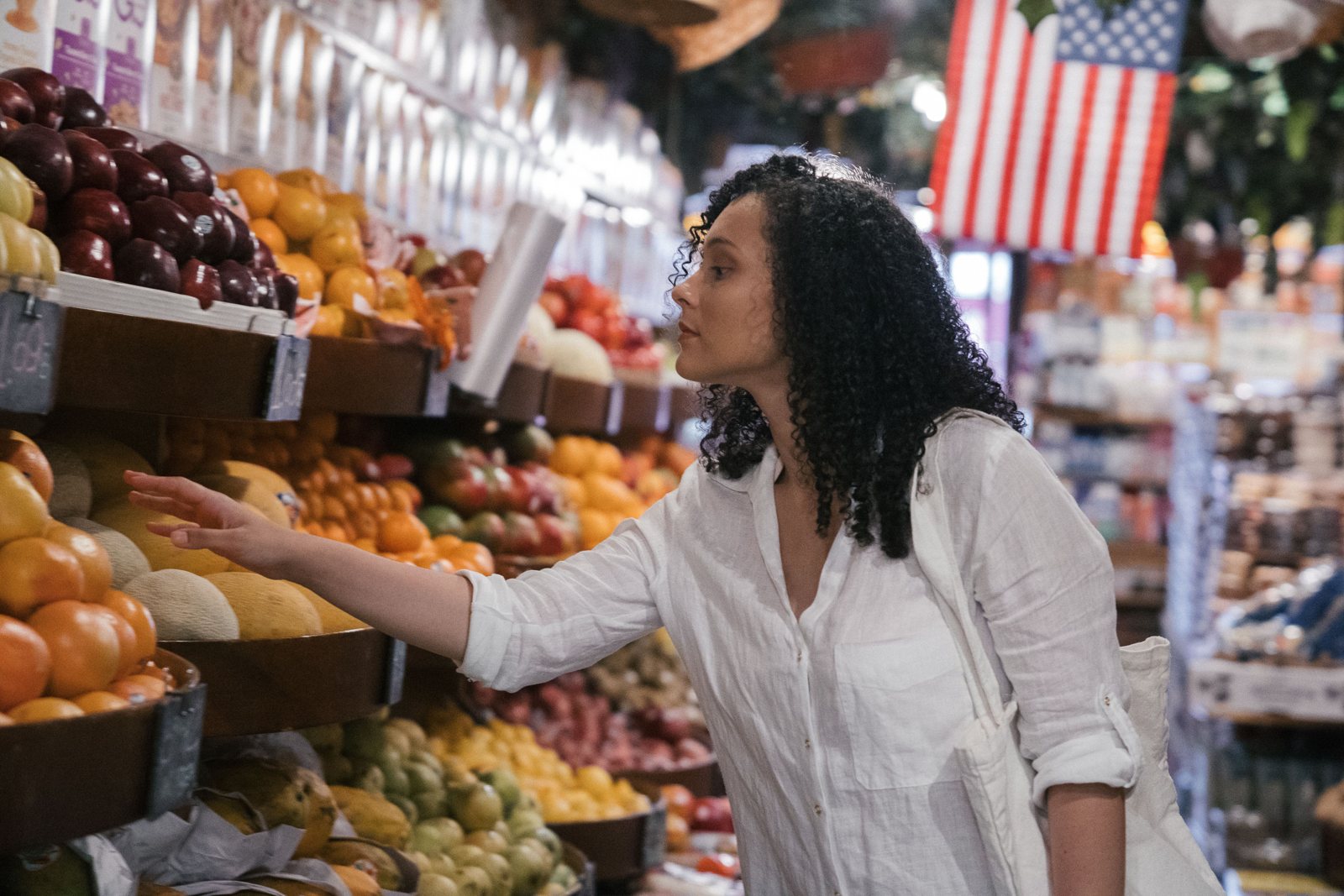 A woman reaching for fresh fruit in a grocery store with an American flag hanging in the background