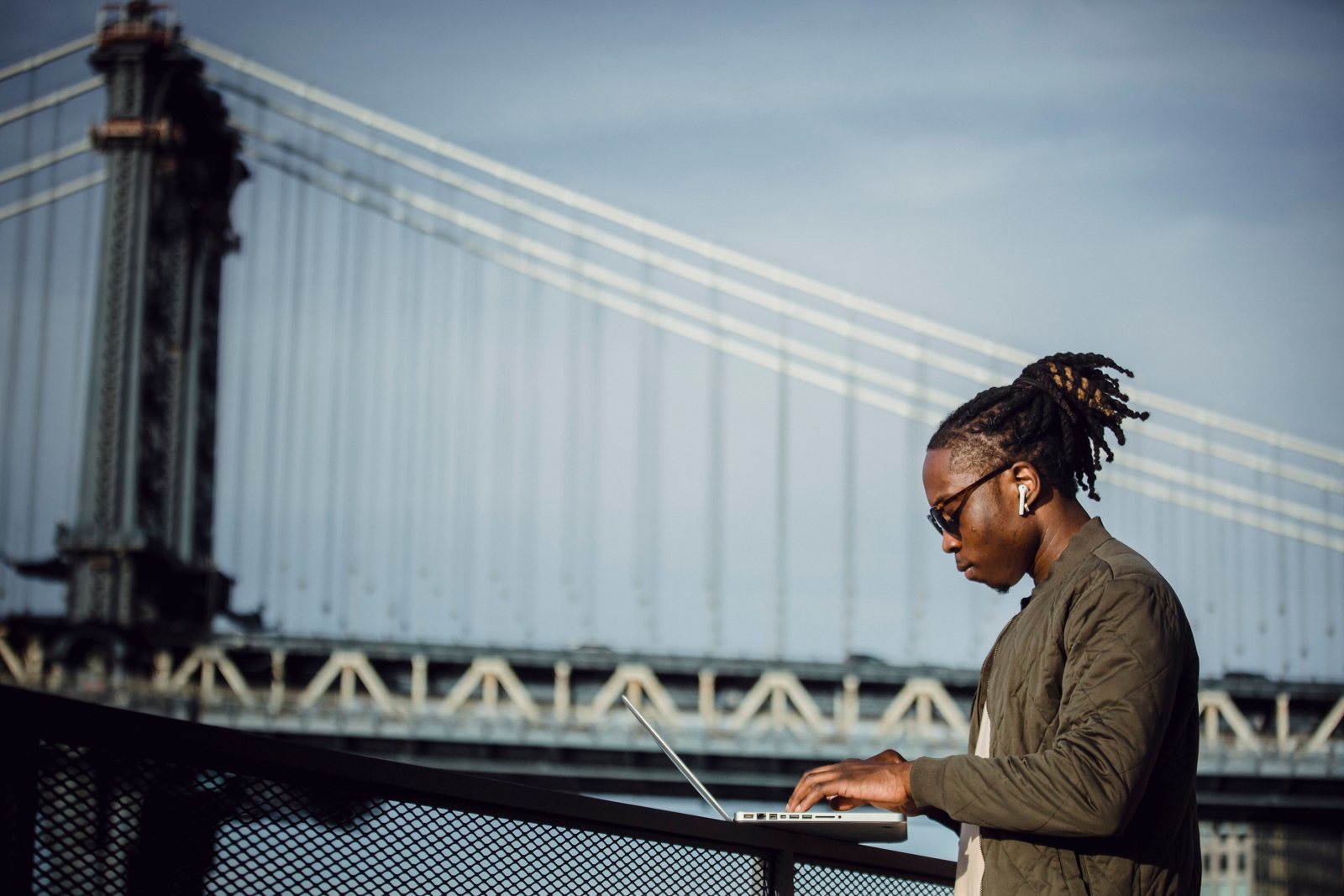 A man in sunglasses typing on a laptop outdoors by the Manhattan Bridge in New York City