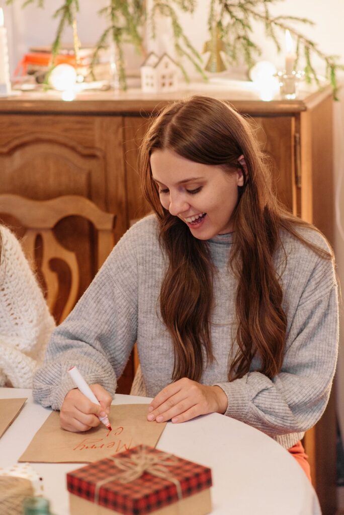A smiling woman in a gray sweater writing on brown paper at a table with a wrapped gift, planning her Christmas sinking fund
