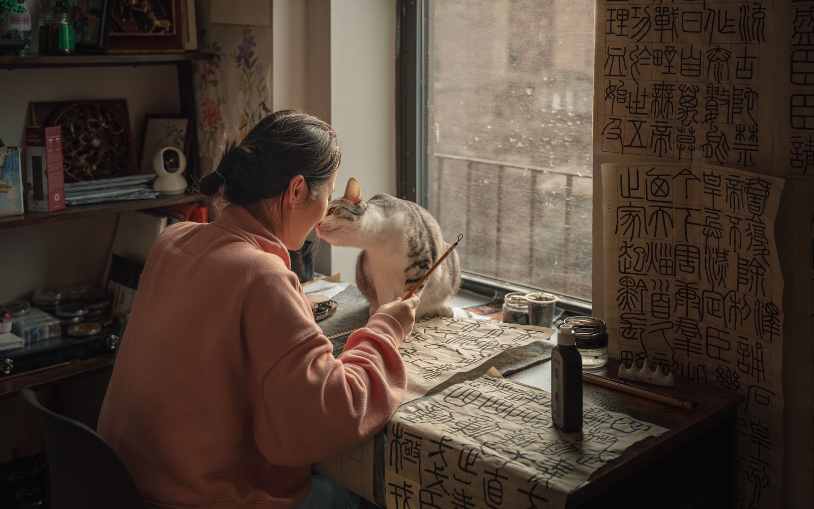 A woman writing by hand at her desk by the window of a New York apartment, her cat beside her