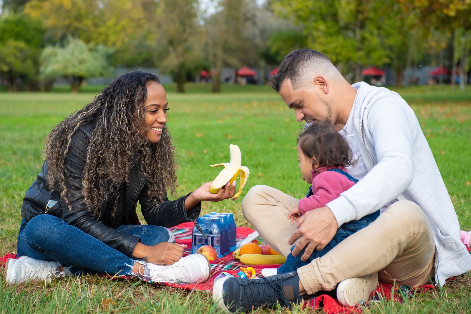 A smiling mother and father sharing fruit and water with their toddler on a red picnic blanket in a grassy park