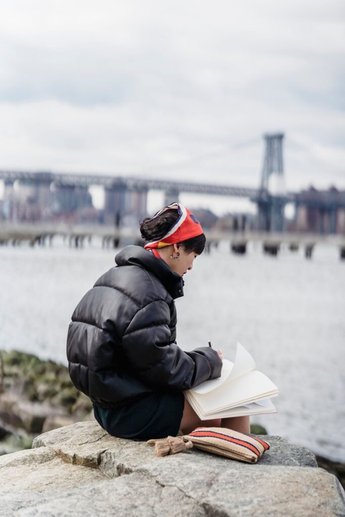 A woman in a warm jacket writing a short plan in a notebook by a New York river, a quiet way to get back on track financially