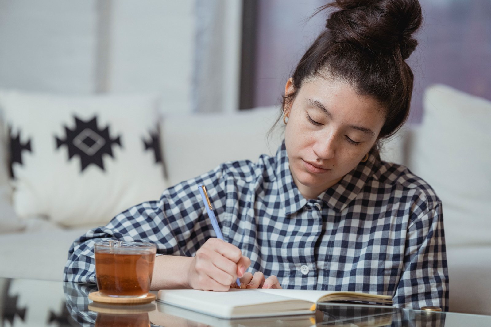 A woman in a checked shirt sitting at home with a cup of tea, writing a simple plan in her notebook to get back on track financially after a rough week
