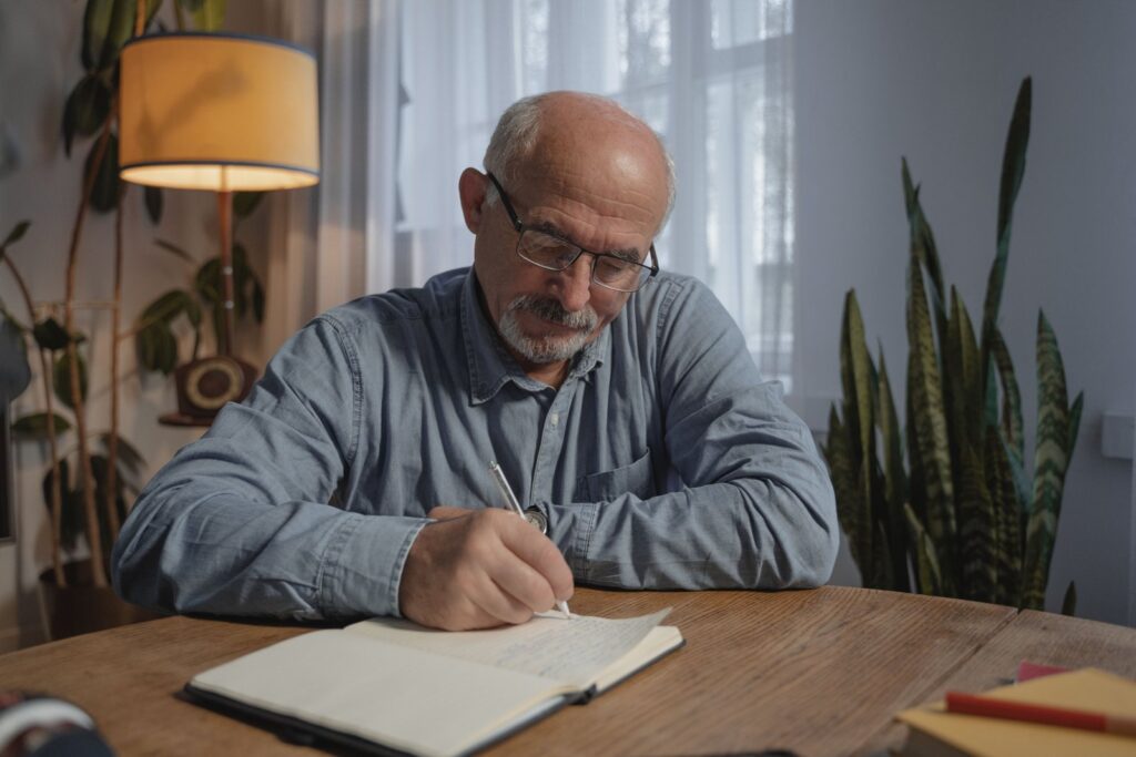 A man in glasses at a wooden table at home, writing notes in a notebook after he calls to request an itemized bill from the hospital