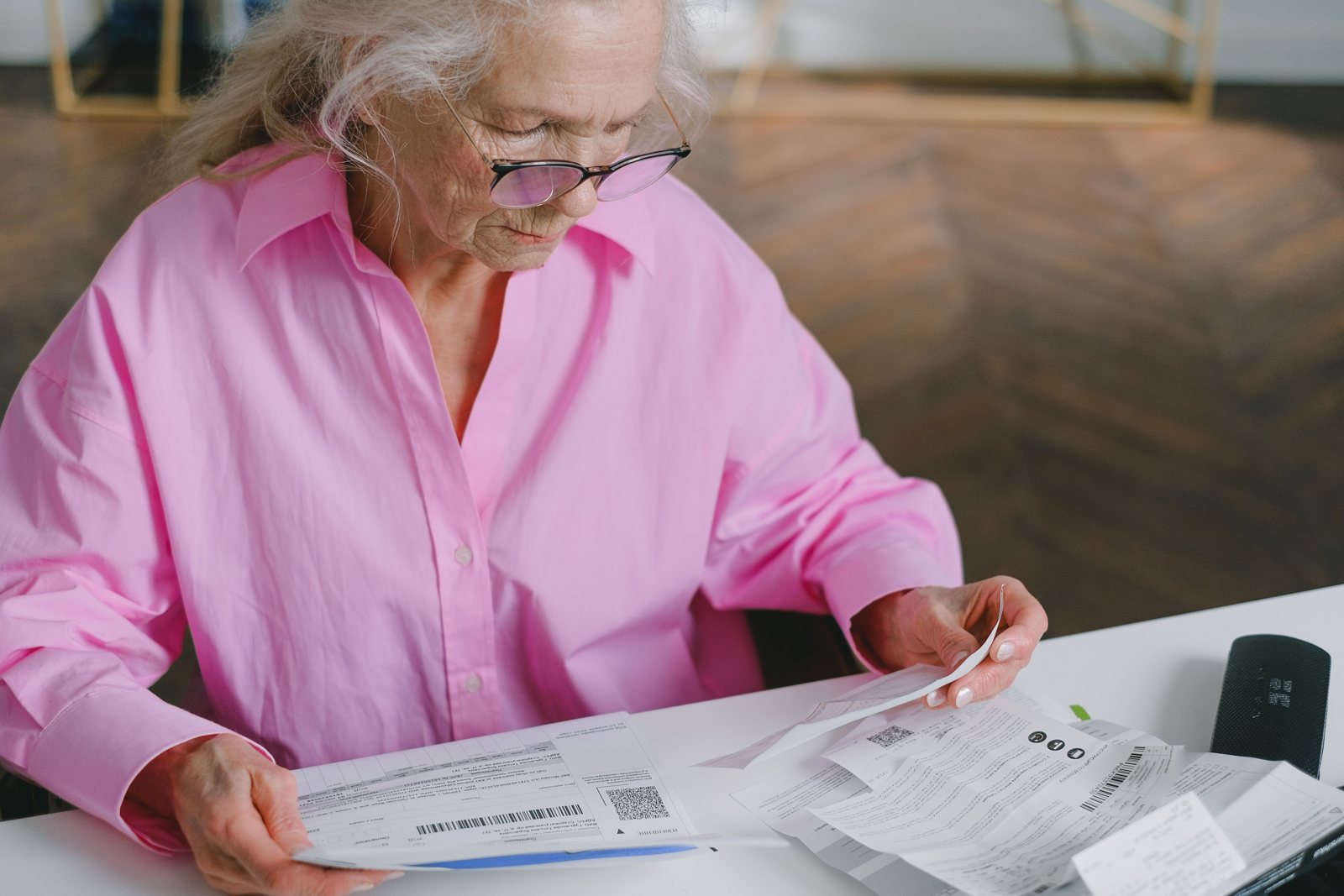 An older woman in a pink shirt and glasses reading through a stack of bills at a table