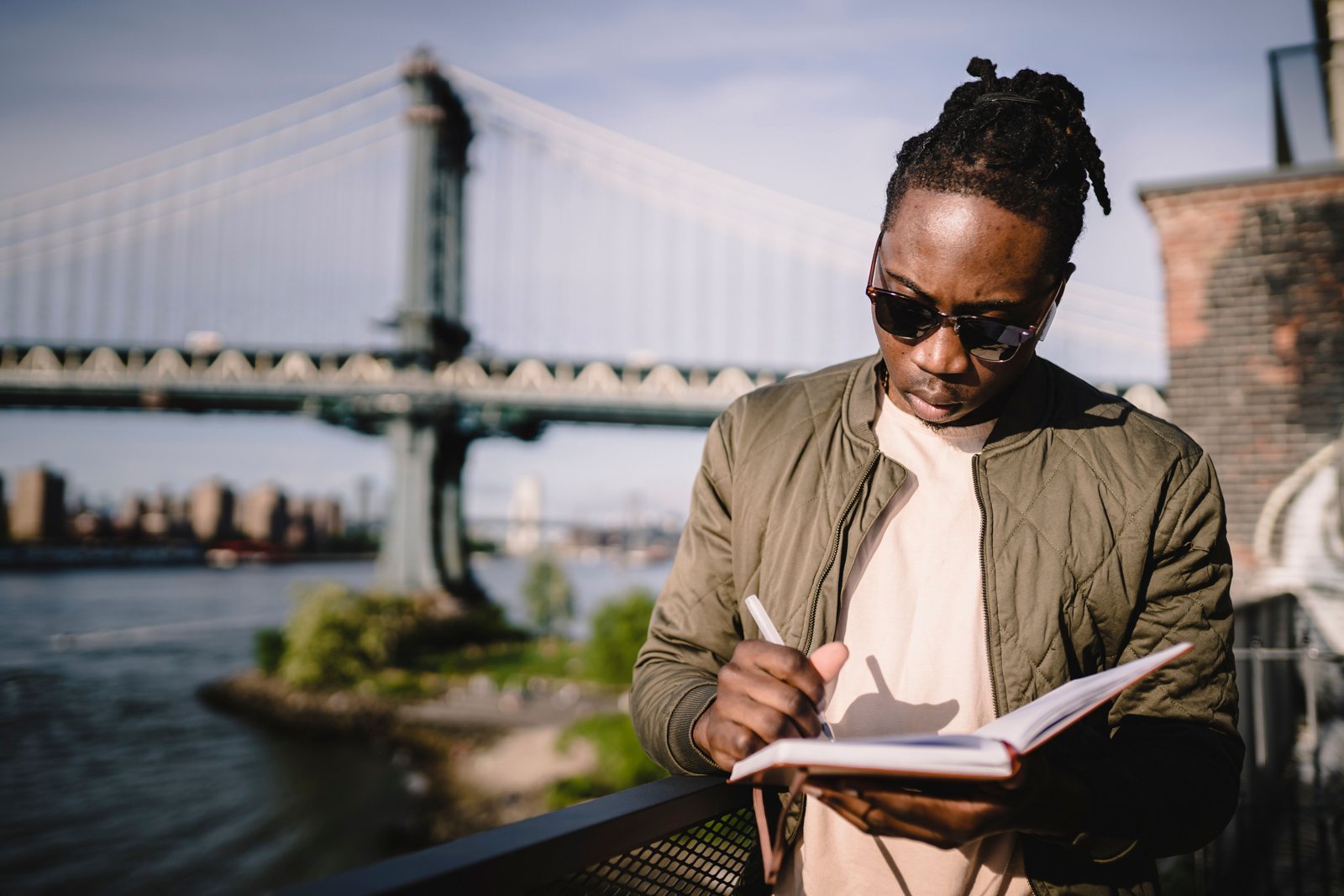 A young man in sunglasses writing in a planner by the river in New York City, with the Manhattan Bridge behind him