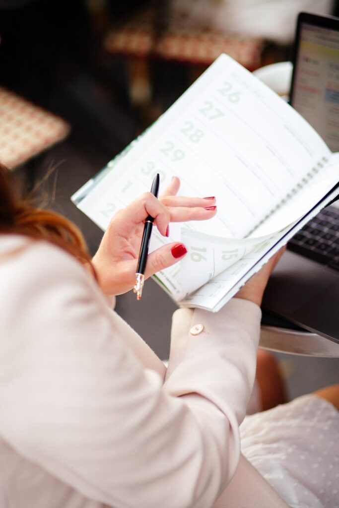 Over-the-shoulder view of a woman with a pen flipping through the dated pages of a paper planner at a cafe table in New York
