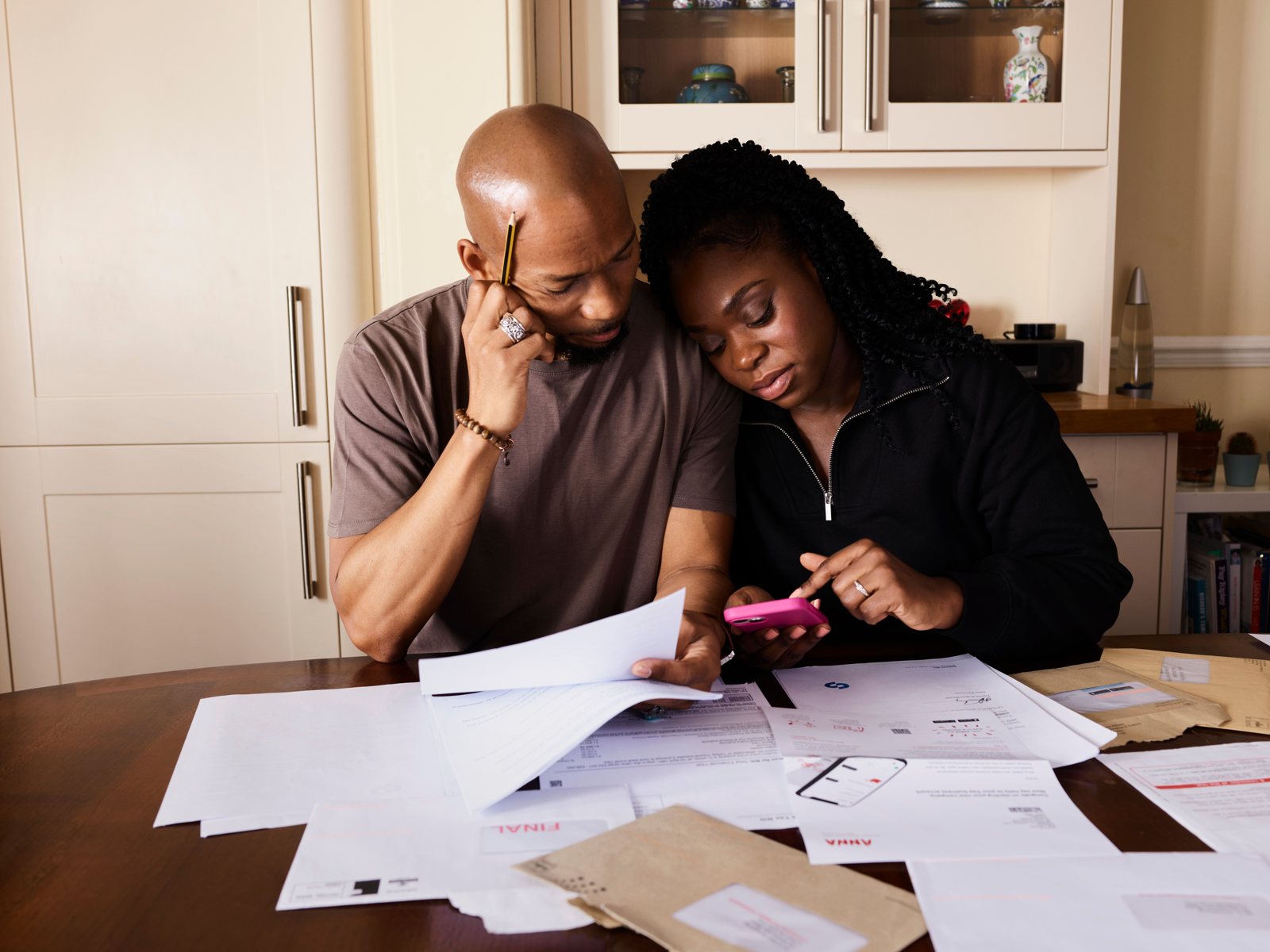 A man and a woman at a kitchen table going over a pile of bills, one holding a phone calculator