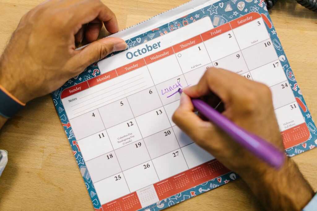 A person writing a note on an October wall calendar with a purple pen while planning the month ahead