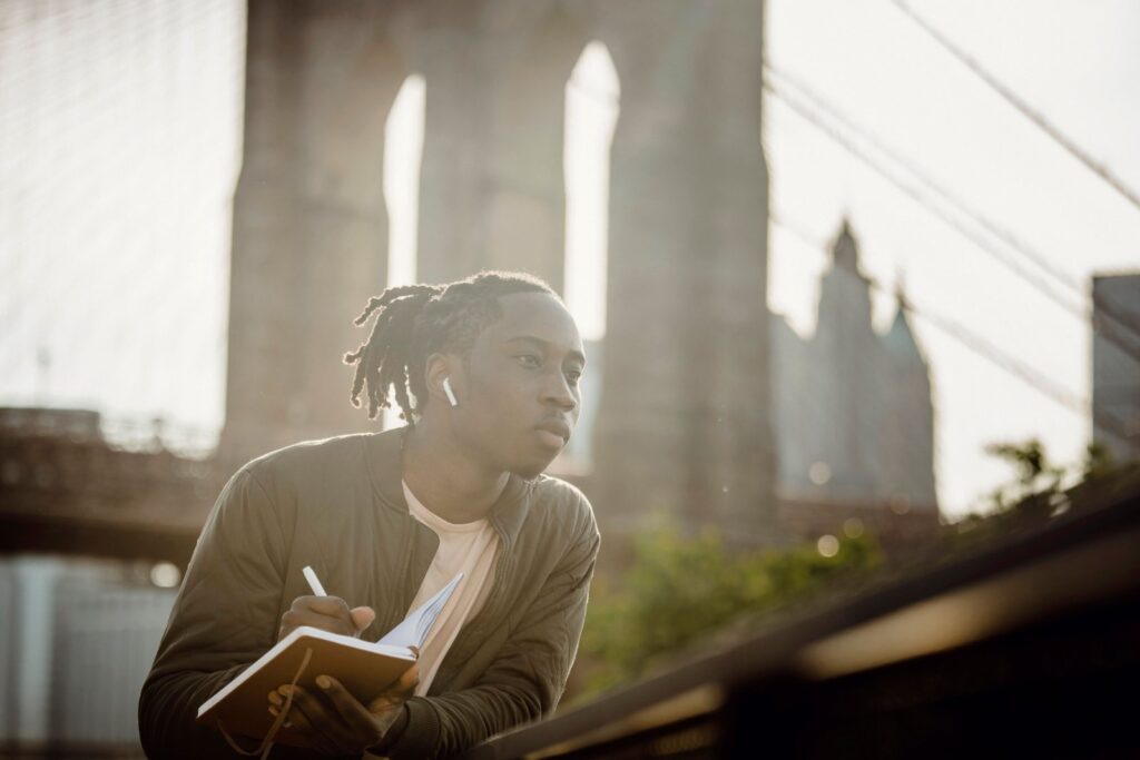 A man writing a short money checklist in a notebook by the Brooklyn Bridge during a weekly money reset