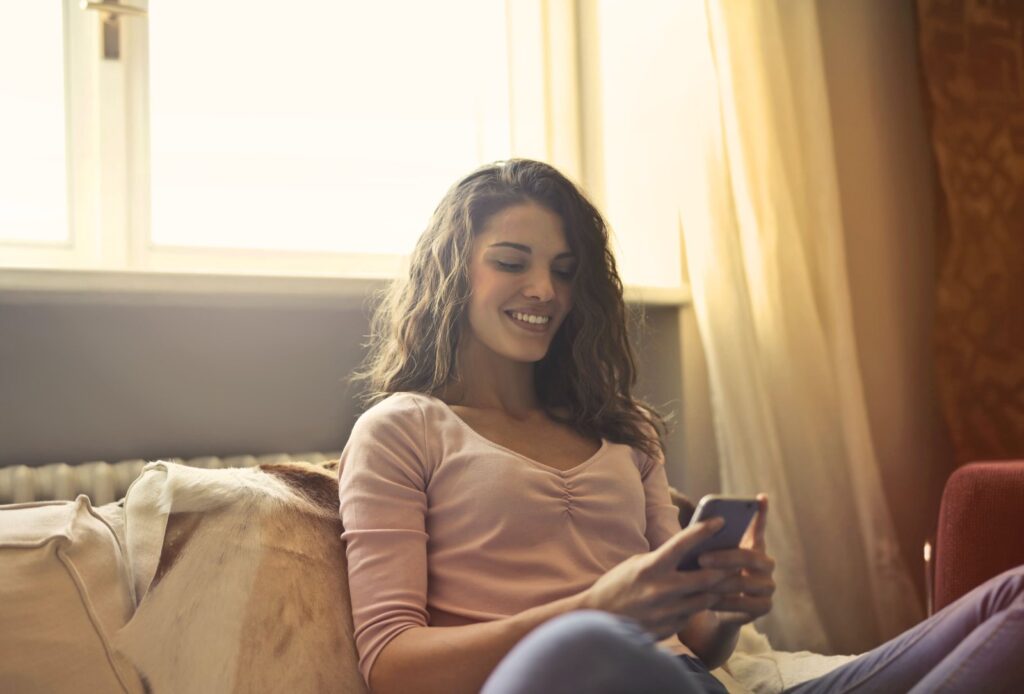 A smiling woman on a couch by a sunny window typing a reply on her phone, a kind way to say no to spending