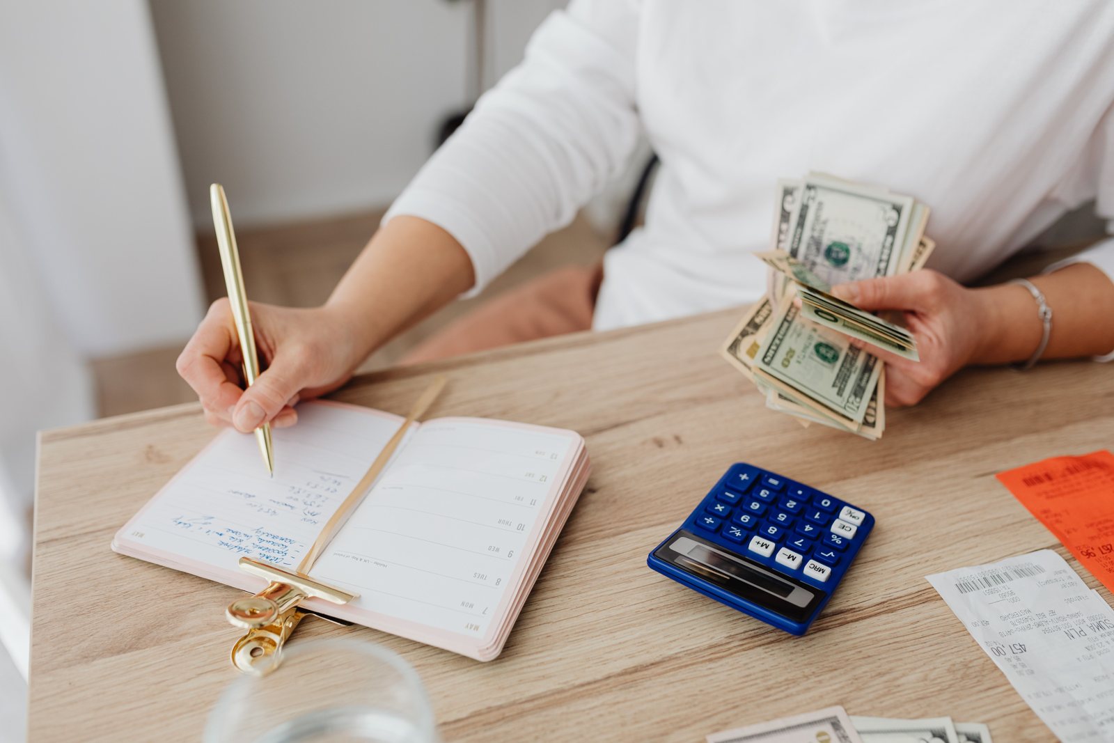A woman holding U.S. dollar bills while writing amounts for her sinking funds in a notebook next to a calculator