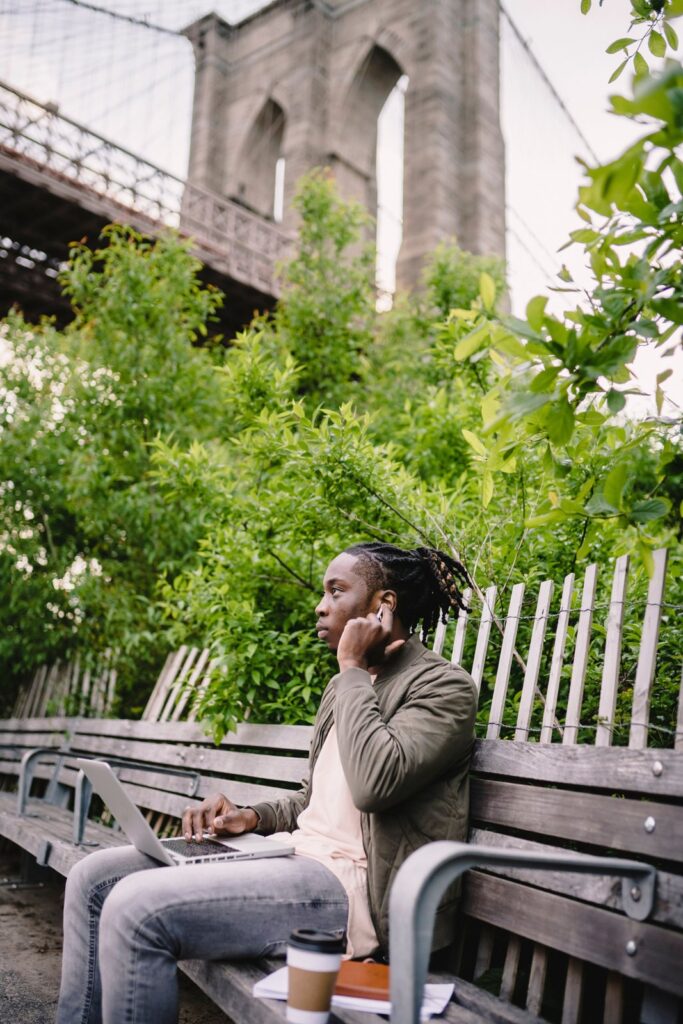 A man on a park bench near the Brooklyn Bridge comparing debt payoff numbers on his laptop
