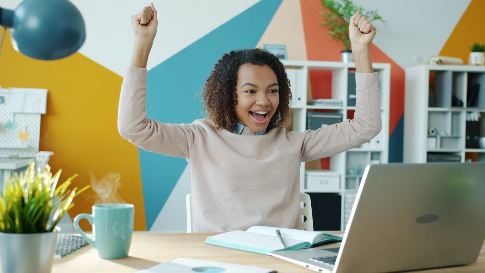 A woman in a sweater raising both fists in celebration at her desk with a laptop and notebook, cheering a small win while paying off debt