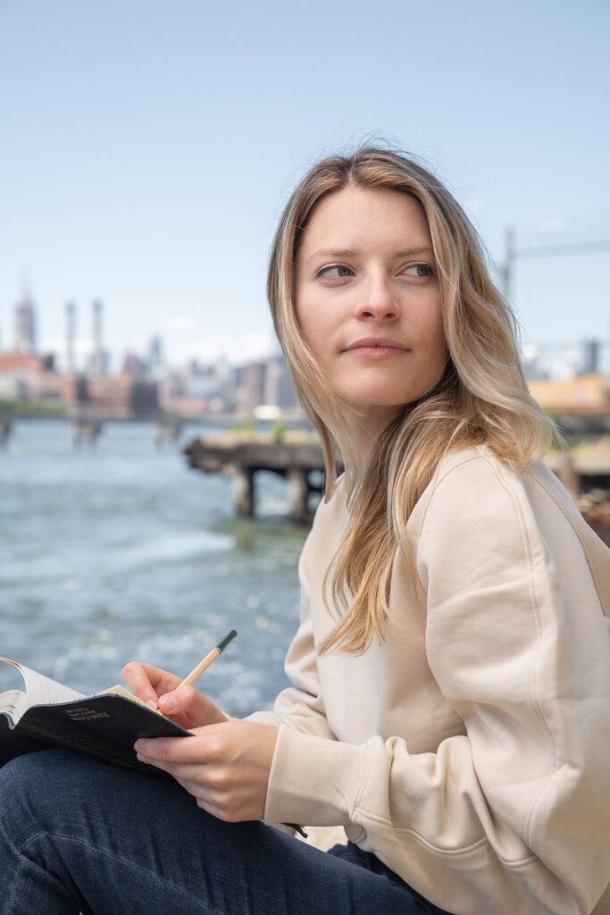 A woman writing in a notebook by the New York waterfront, staying motivated paying off debt with a weekly win log