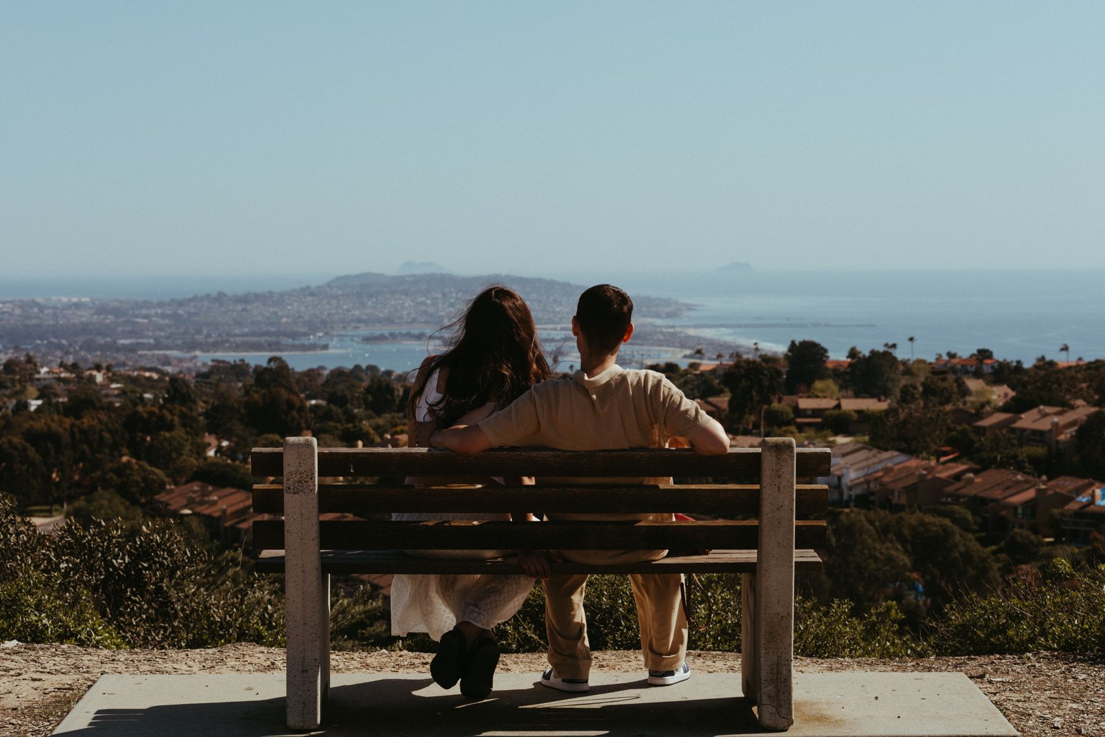 A couple sitting together on a hilltop bench in San Diego, looking out over the city and the coastline