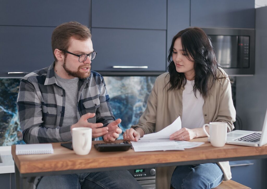 A husband and wife sitting at their kitchen table at home, calmly going over bills and papers together with coffee, a laptop and a calculator