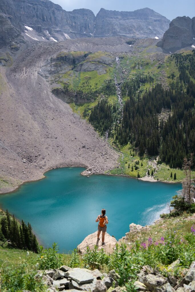 A hiker standing on a rocky overlook above a turquoise alpine lake in the United States, looking out toward mountain peaks