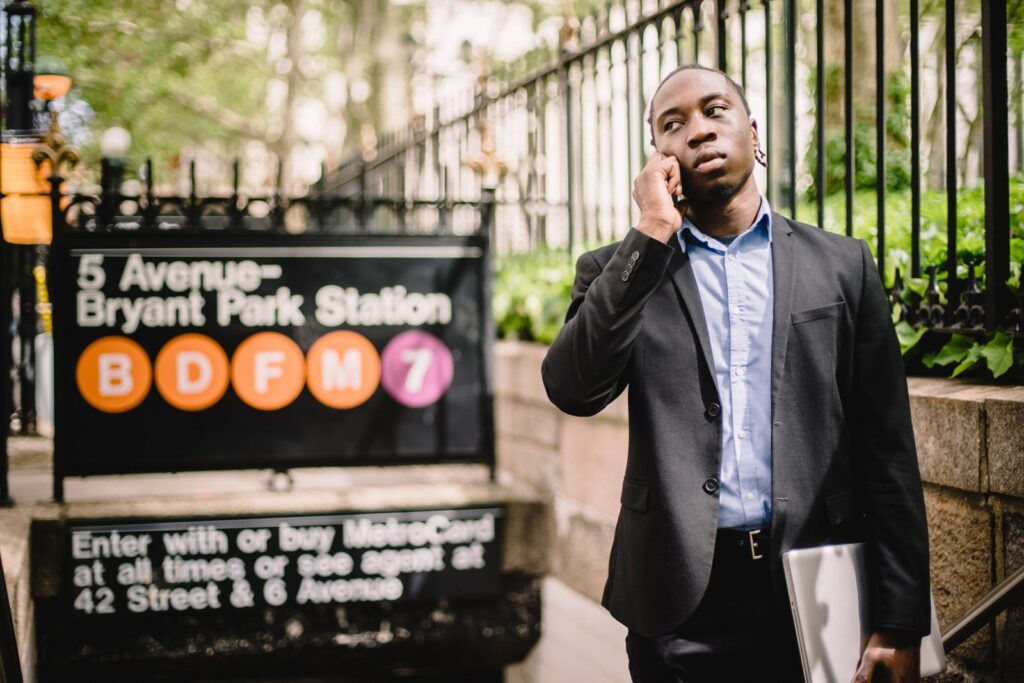 A man near a New York City subway entrance talking on his phone while holding a laptop under his arm