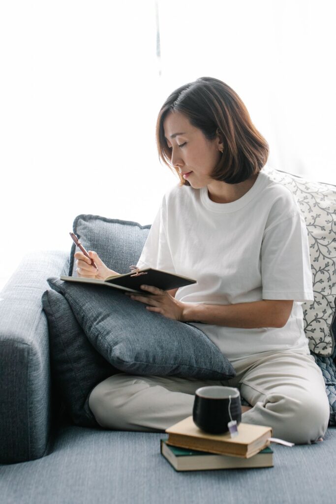 A woman on a couch writing her money story in a small journal, with a cup of tea beside her